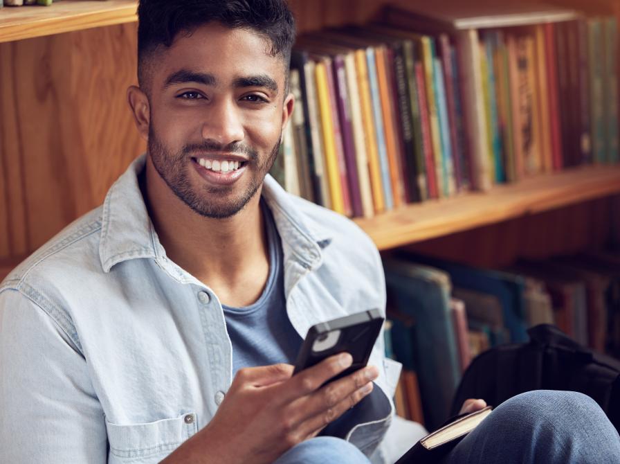 Mand sidder ved reol med bøger med sin mobiltelefon